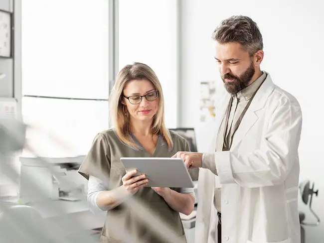 Ein Arzt in weißem Kittel zeigt einer Frau in medizinischer Kleidung Informationen auf einem Tablet in einer hellen Praxis.